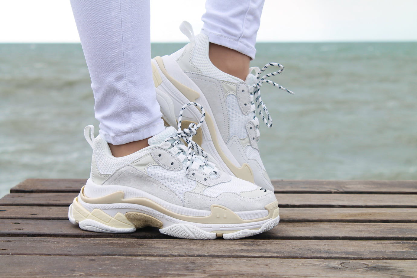 A person wearing chunky off-white and beige chunky sneakers with white-and-black patterned laces, standing on a wooden dock with the ocean in the background.