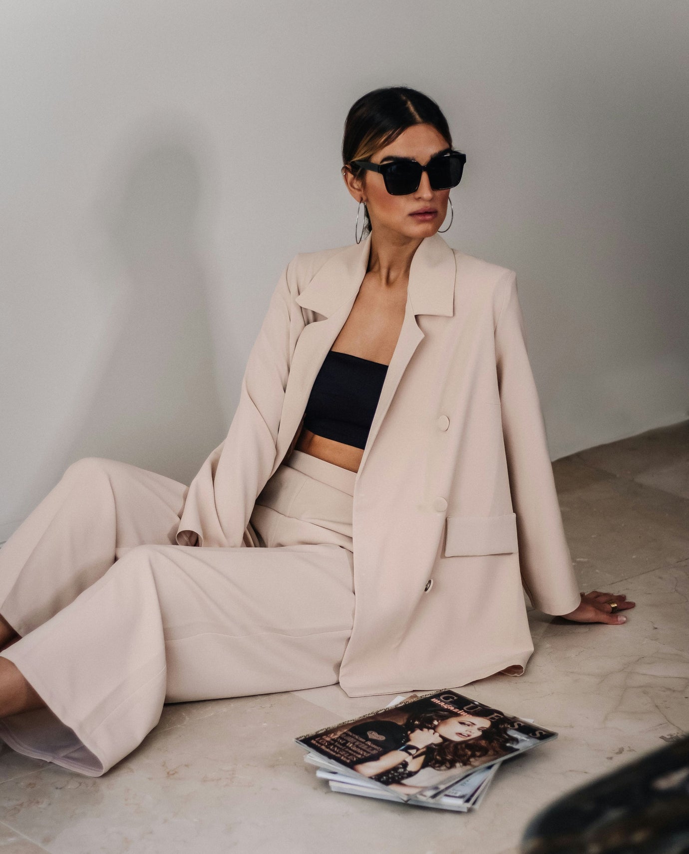 A woman wearing a beige double-breasted blazer and matching wide-leg trousers sits on a light stone floor against a neutral wall, styled with a black bandeau top and black sunglasses.
