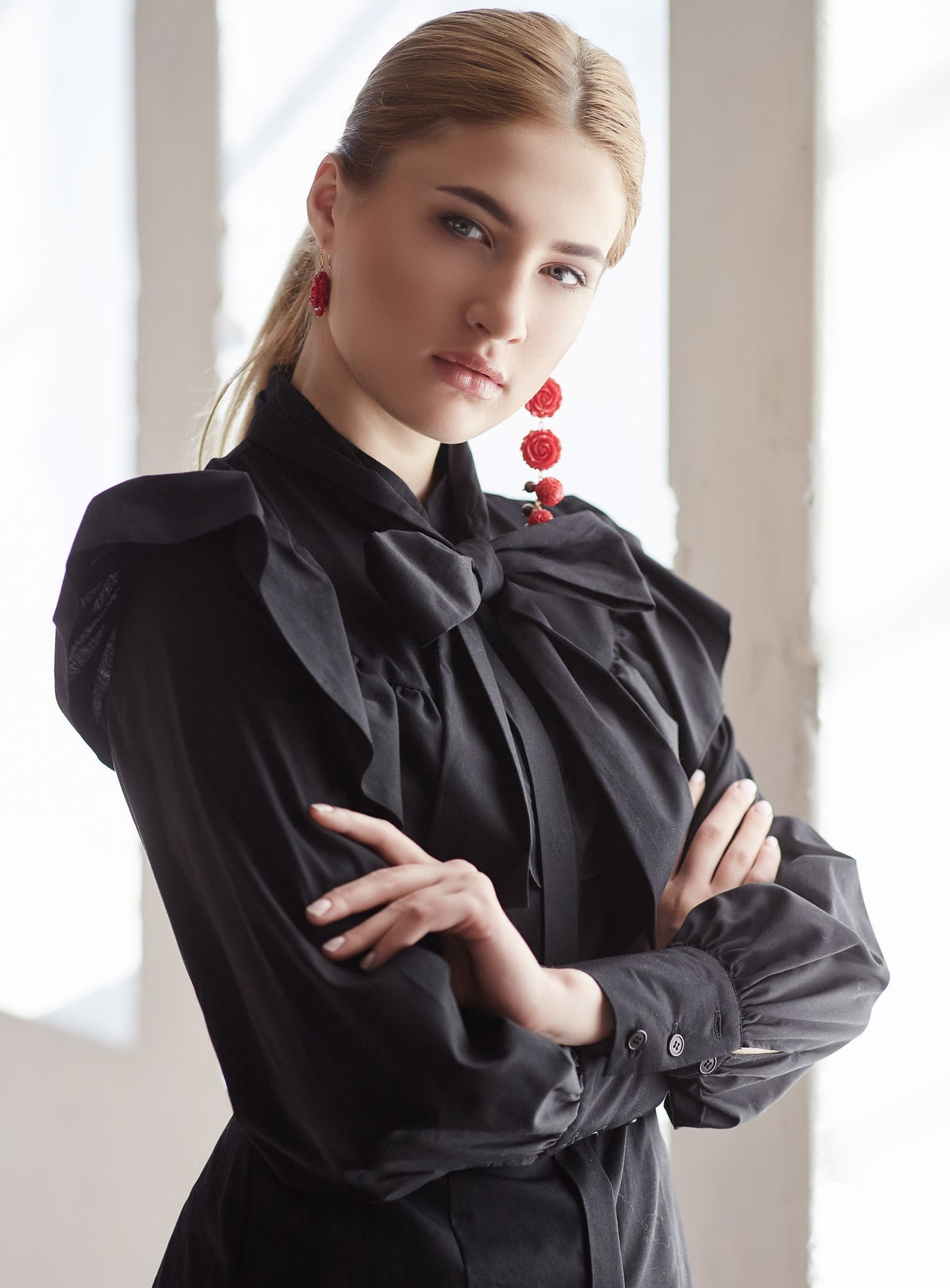 A woman with blonde hair tied back wears a black long-sleeve blouse with dramatic ruffles and a large bow at the collar, paired with red statement earrings, posed with arms crossed against a bright, softly lit window background.