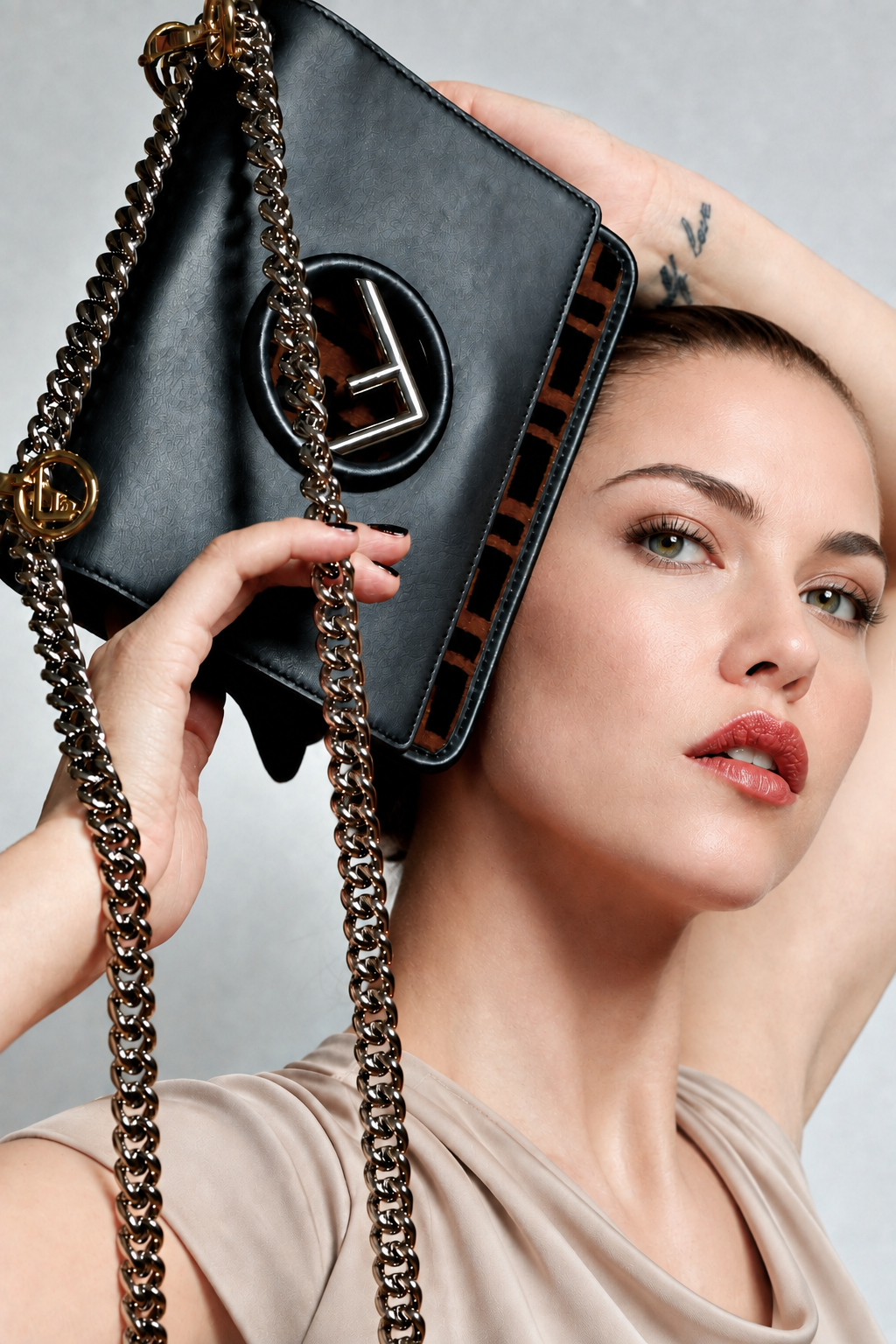 A close-up lifestyle shot of a model holding a black leather shoulder bag with a gold-tone chain strap and a prominent circular gold logo on the front, set against a neutral gray background.